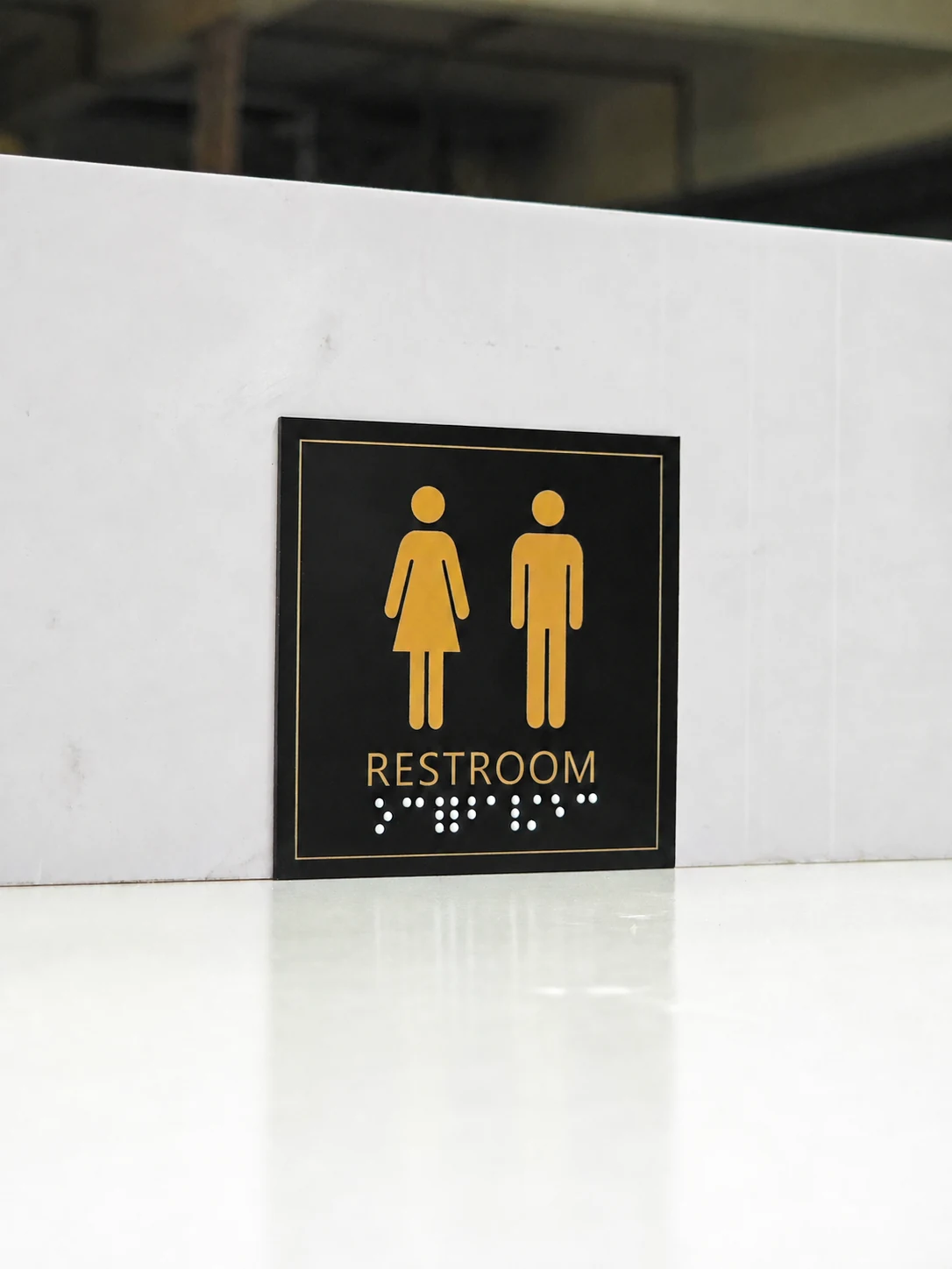Black and Gold Resin Braille Restroom Sign — Full front view showing the black-and-gold layout and two groups of white tactile dots below the RESTROOM text.