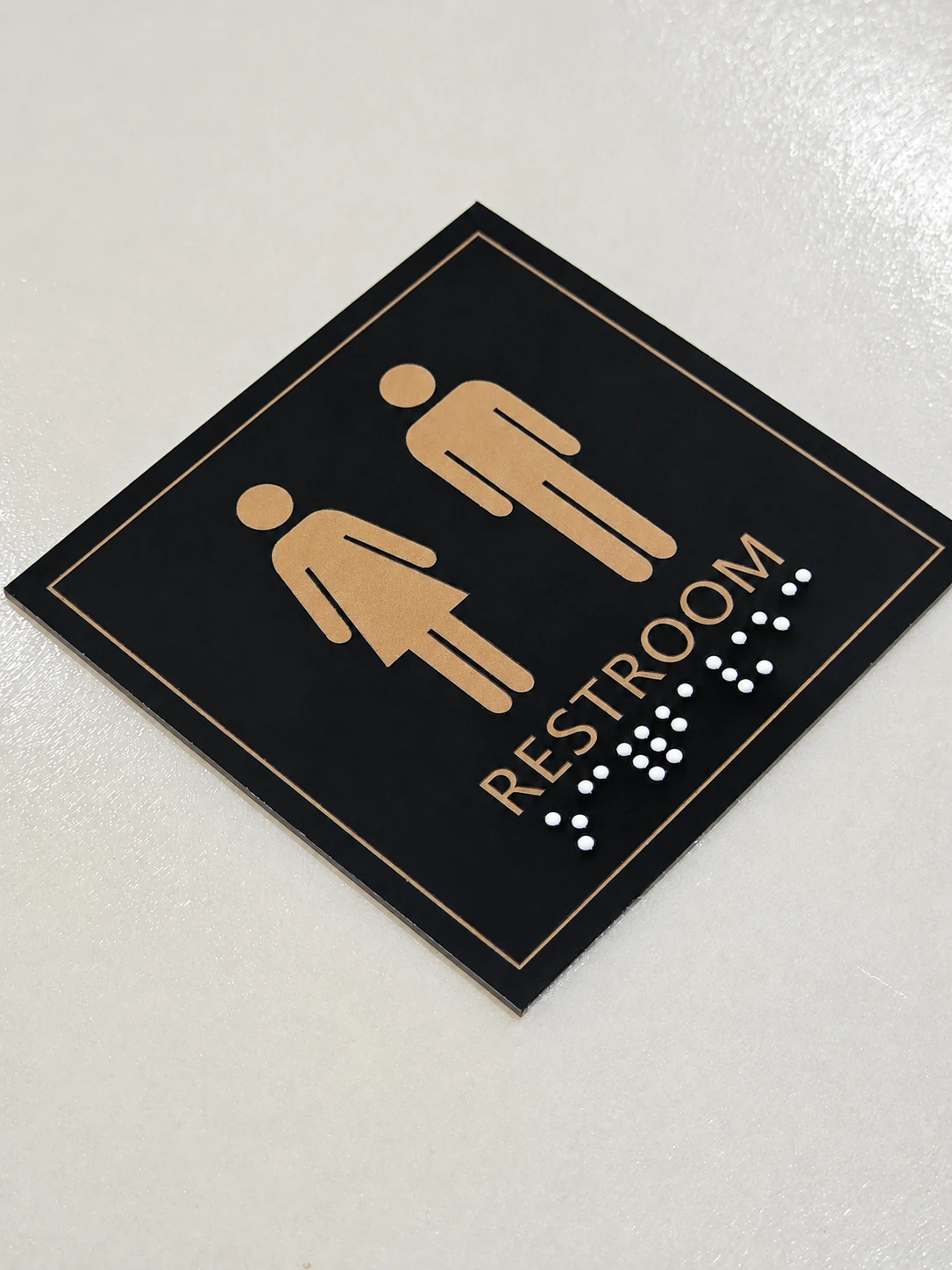 Black and Gold Resin Braille Restroom Sign — Angled full view showing the raised gold icons and text, white dots, square edge, and surrounding border.